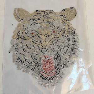 May include: A close-up of a tiger face made of rhinestones. The tiger's face is white with black stripes and has red eyes. The rhinestones are in various colors, including black, white, gold, and red.