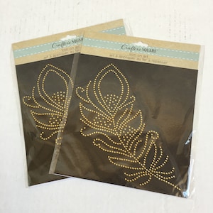 May include: Two packages of iron-on art featuring a gold peacock feather design. The packages are labeled "Crafter's Square" and "Iron-on art".