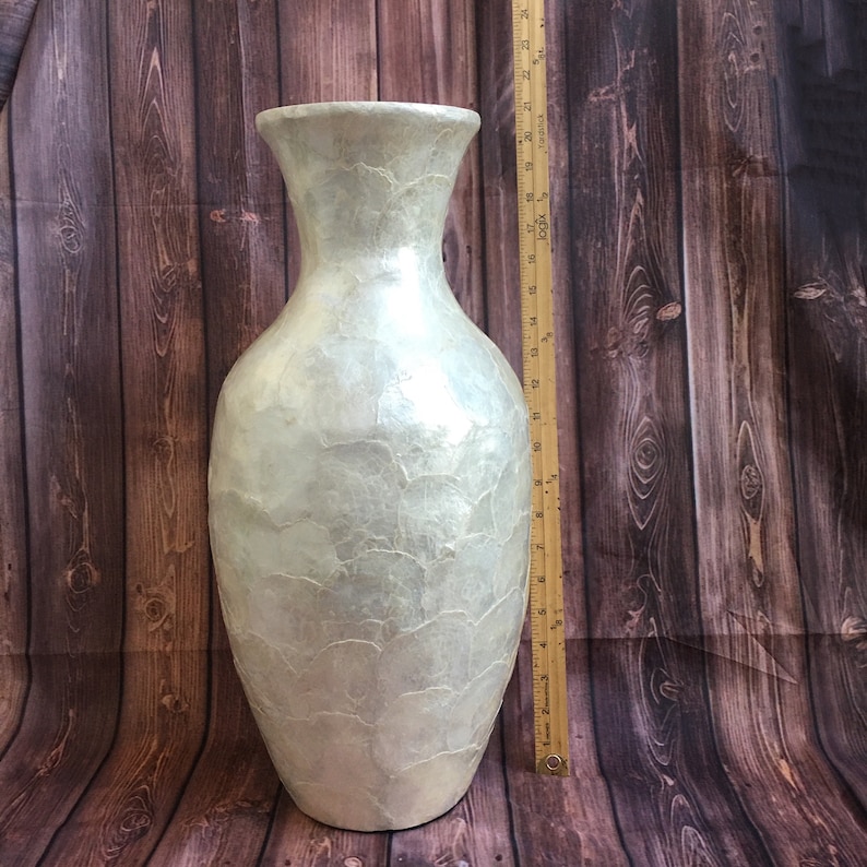 May include: A tall, pearlescent white vase with a textured surface. The vase has a wide base, a slender neck, and a flared opening. The vase is approximately 24 inches tall.