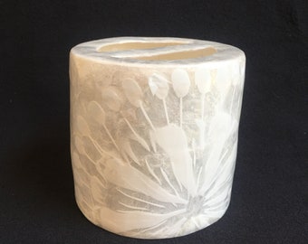 May include: White ceramic cylindrical vase with a raised floral design. The vase has a smooth, textured surface and a slightly rounded top.
