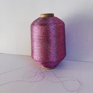 May include: A spool of pink metallic thread with a wooden center. The thread is unwinding from the spool.