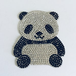 May include: A sparkly panda bear-shaped embellishment. The panda's body is covered in clear, iridescent rhinestones, with black rhinestones outlining the ears, eyes, nose, and limbs. The design is set against a white background.