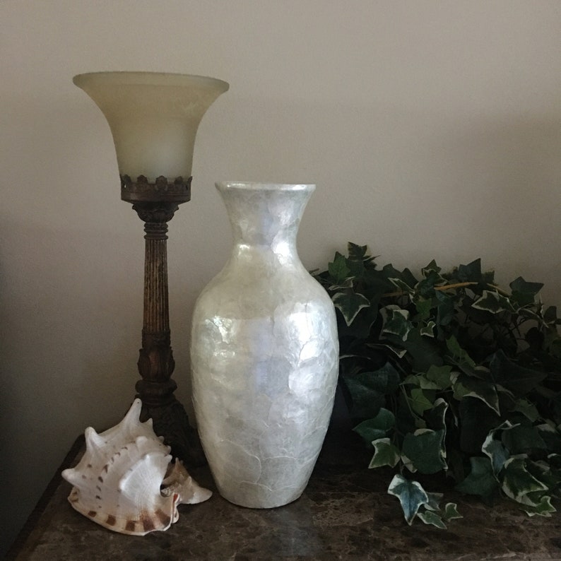 May include: A pearlescent white vase with a textured surface stands next to a vintage lamp with a frosted glass shade. A large seashell with brown and white markings rests on the table, with green foliage in the background. Home decor.