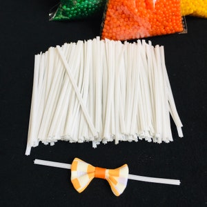 May include: A large quantity of white twist ties, also known as zip ties, are arranged in a pile. A single twist tie is tied into a bow shape, with the ends of the tie extending out to the sides.