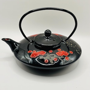 May include: Black ceramic teapot with a red floral design and a black handle. The teapot has a lid with a small hole in the top.
