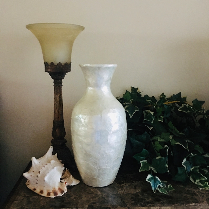 May include: A pearlescent white vase with a textured surface stands next to a vintage lamp with a frosted glass shade. A large seashell with brown and white markings rests on the table, with green foliage in the background.