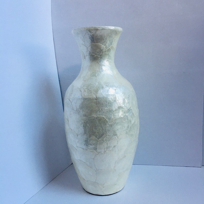 May include: A tall, pearlescent white vase with a textured surface. The vase has a wide base that tapers to a slender neck and flared opening. The surface has a cracked, mosaic-like appearance. This decorative vase is suitable for floral arrangements.