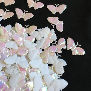 May include: A large pile of iridescent white and pink butterfly shaped embellishments. The butterflies are made of a shiny, pearlescent material.