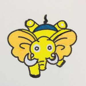May include: A yellow sequined elephant applique with black beaded outlines. The elephant has large, fan-shaped ears, white tusks, and a blue and yellow head. The applique is on a white background.