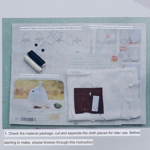 May include: A white fabric kit for making a Totoro plushie. The kit includes fabric pieces, thread, buttons, and instructions. The instructions include a diagram of the Totoro's body parts and a list of materials. The text on the instructions reads "Mengwuchi My Neighbor Totoro Series Ornament My Neighbor Totoro Material Pack List" and "Kitty Totoro Material Kit for Cat Series Ornament Production Instructions".