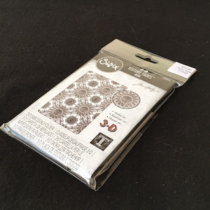 May include: A close-up of a 3-D Texture Fades Mini embossing folder by Sizzix. The folder is packaged in a clear plastic bag with a white and black label. The label features the Sizzix logo, the product name, and the price. The folder is designed to create a 3-D effect on paper or cardstock.