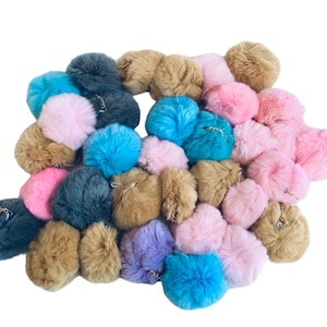 May include: A collection of fluffy pom-pom keychains in various colors, including pink, blue, brown, and gray. The keychains are made of soft, fuzzy material and have a gold chain attached.