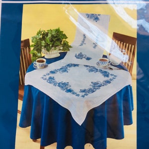 May include: White tablecloth with a blue embroidered border of teapots and flowers. The tablecloth is laid on a blue table and there are two cups and saucers on the table.