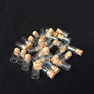 Puede incluir: Un grupo de peque&ntilde;as botellas de vidrio transparente con tapones de corcho. Las botellas est&aacute;n dispuestas en una pila sobre una superficie negra.