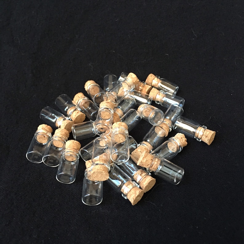 Puede incluir: Un grupo de peque&ntilde;as botellas de vidrio transparente con tapones de corcho. Las botellas est&aacute;n dispuestas en una pila sobre una superficie negra.