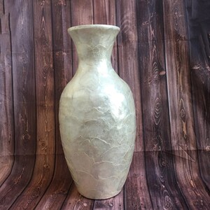 May include: A tall, pearlescent vase with a wide base and flared opening. The vase has a textured surface with a subtle, crackled pattern. The color is a soft, muted white with hints of silver, creating a shimmering effect. The vase is a decorative home accent.