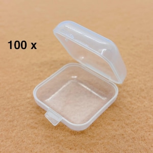 May include: A clear, square plastic container with a hinged lid, open to reveal the interior. The container is white and has a small latch. The text "100 x" is printed in black on the left side. The background is a light brown.