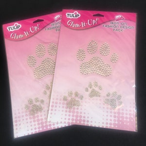 May include: Three packs of iron-on fashion design featuring paw prints with rhinestones. The packs are pink and white with the text "TULIP Glam-It-Up!" and "IRON-ON FASHION DESIGN PACK".