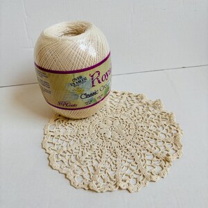 May include: A ball of off-white crochet thread next to a crocheted doily. The thread is labeled "Royal Classic Crochet" and wrapped in plastic. The doily is circular with a detailed, openwork design.