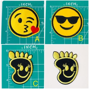 May include: Four embroidered iron-on patches featuring yellow smiley faces with different expressions. The first patch shows a winking smiley face with a red heart. The second patch shows a smiley face wearing sunglasses. The third and fourth patches show a smiley face with closed eyes and eyelashes. The patches are approximately 2 inches in diameter.