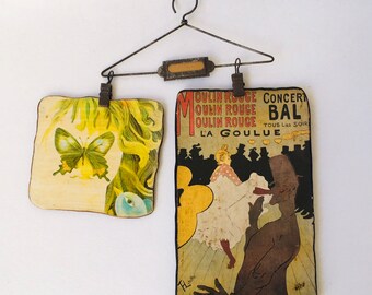 May include: Two vintage postcards hanging on a wire hanger. The left postcard features a butterfly and a woman's face. The right postcard is a Moulin Rouge poster with the text "Concert Bal Moulin Rouge Tous Les Soir La Goulue".