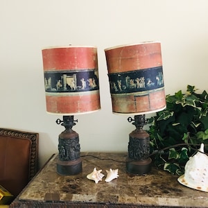 May include: Two vintage lamps with red and black patterned lampshades. The lamps have a bronze base with a detailed design. The lamps are on a brown table with two seashells.