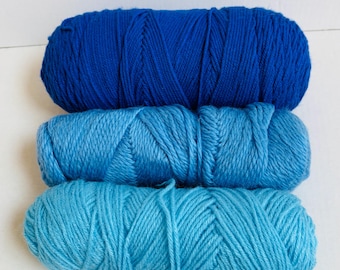 May include: Three skeins of yarn in shades of blue are stacked on top of each other. The top skein is a deep blue, the middle is a medium blue, and the bottom is a light blue. The yarn is tightly wound and appears soft.