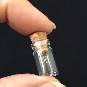 Puede incluir: Una peque&ntilde;a botella de vidrio transparente con un tap&oacute;n de corcho. La botella mide aproximadamente 5 cm de alto y tiene un di&aacute;metro de unos 2,5 cm.