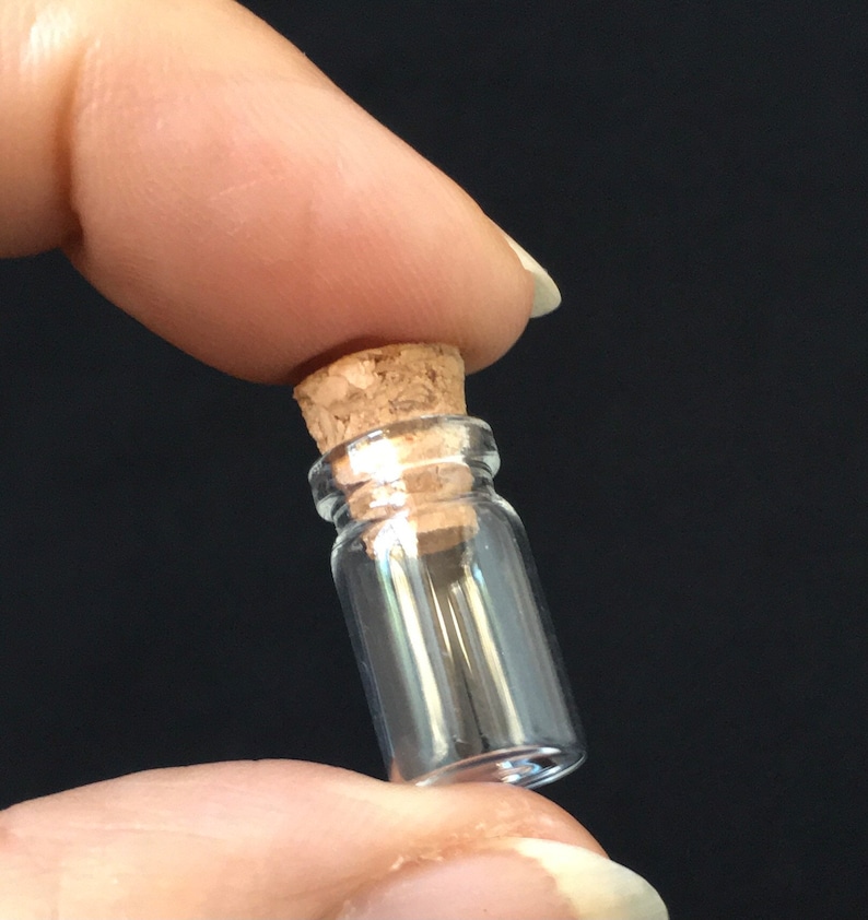 Puede incluir: Una peque&ntilde;a botella de vidrio transparente con un tap&oacute;n de corcho. La botella mide aproximadamente 5 cm de alto y tiene un di&aacute;metro de unos 2,5 cm.