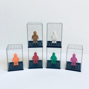 May include: Six clear plastic display cases with black bases, each containing a small Lego minifigure. The minifigures are in various colors, including orange, brown, red, green, pink, and white.