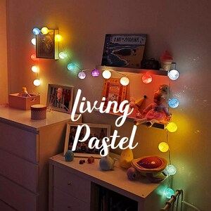 Könnte beinhalten: Ein warmer, einladender Raum mit Lichterketten, die einen Raum beleuchten. Die bunten, kugelförmigen Lichter sind über einer weißen Kommode und einem Regal drapiert. Der Text "living Pastel" ist in einer Schreibschrift dargestellt.