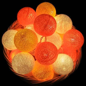 Puede incluir: Una cadena de luces de bola de algodón en una cesta de mimbre. Las luces son en tonos de naranja, amarillo y blanco.