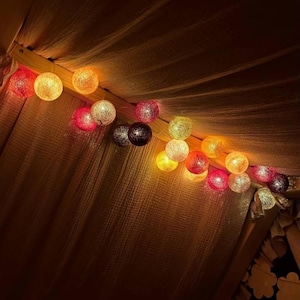 May include: A string of spherical lights featuring a textured, woven surface. The globes emit a warm glow in various colors, including shades of red, orange, yellow, and white, arranged in a linear sequence along a thin, flexible electrical cord.