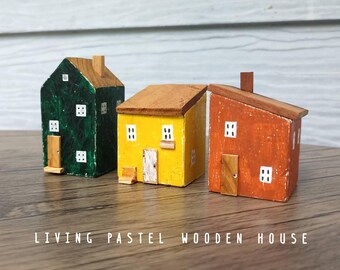 Puede incluir: Tres pequeñas figuras de casas de madera en verde, amarillo y naranja con techos marrones. Cada casa presenta ventanas blancas pintadas y una pequeña puerta de madera. El texto "LIVING PASTEL WOODEN HOUSE" aparece impreso en blanco.