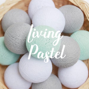 Può includere: Un cesto pieno di morbide palle di cotone rotonde in tonalità di grigio, bianco e verde chiaro. Il testo "living pastel" è sovrapposto all'immagine.