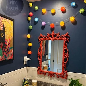 Pode incluir: Um fio de luzes LED esféricas multicoloridas em tons de amarelo, laranja, vermelho, verde, azul e roxo pendurado numa parede azul-escura sobre um espelho retangular com uma moldura vermelha ornamentada e um revestimento de azulejos brancos.