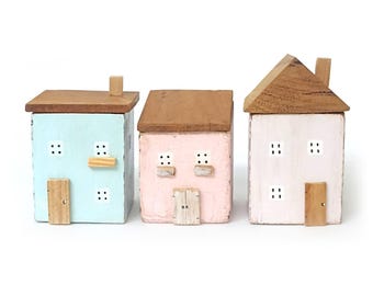 Può includere: Tre piccole statuette di case in legno disposte in fila. La casa a sinistra è azzurra, quella centrale è rosa chiaro e quella a destra è bianco sporco. Ognuna presenta un tetto in legno marrone, una piccola porta e finestre bianche dipinte.