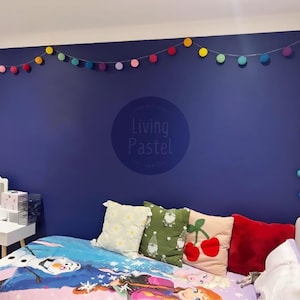 Pode incluir: Interior de quarto com uma parede azul escuro adornada com uma guirlanda de luzes de pompons coloridos. Uma cama é coberta com uma colcha estampada e almofadas variadas. Uma penteadeira branca com espelho também é visível.