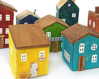 Puede incluir: Una colección de pequeñas figuras de casas de madera pintadas a mano en varios colores, incluyendo amarillo, turquesa, verde, rojo, azul y naranja. Cada edificio en miniatura presenta un techo marrón, ventanas blancas pintadas y una pequeña puerta.