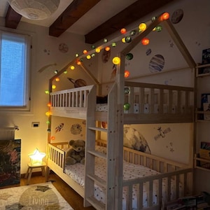 May include: A wooden bunk bed with a house-shaped frame, decorated with colorful string lights. The room features space-themed wall decals, a star-shaped lamp, and a teddy bear on the lower bunk. The room is decorated with a rug and a painting.