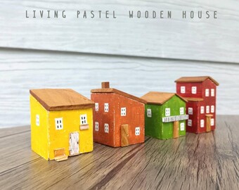 Puede incluir: Cuatro pequeñas figuras de casas de madera dispuestas en fila. Las casas están pintadas de amarillo, naranja, verde y rojo, cada una con un techo inclinado marrón, ventanas pintadas de blanco y una pequeña puerta. El texto LIVING PASTEL WOODEN HOUSE es visible.