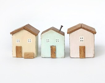 Può includere: Tre piccole statuette di case in legno con tetti marroni. La casa a sinistra è color crema con un piccolo gradino, quella centrale è verde chiaro con un camino in metallo e quella a destra è rosa pallido. Ognuna presenta una porta in legno.