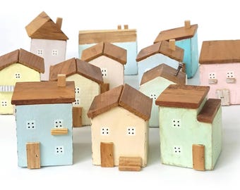 Puede incluir: Una colección de pequeñas figuras de casas de madera pintadas a mano en varios colores pastel, incluyendo azul claro, amarillo pálido, verde menta y rosa suave. Cada edificio en miniatura tiene un techo de madera marrón, ventanas y puertas.