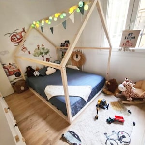 May include: A wooden house-shaped bed frame with a blue mattress, white blanket, and string lights. The room features wall decals of a helicopter and truck, a rug with vehicle illustrations, and a sign hanging by the window reading MY ROOM MY RULES.