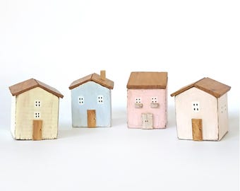 Può includere: Un set di quattro piccole statuette di case in legno con tetti a capanna. Le case sono dipinte in giallo pastello, azzurro, rosa tenue e bianco sporco. Ogni casa presenta una piccola porta in legno, finestre dipinte e un tetto color legno.