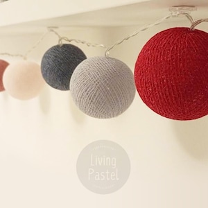 May include: A string of decorative globe lights in various colors, including red, pink, and gray. The lights are made of a woven material and are strung together on a clear wire. The lights are hanging from a white shelf.