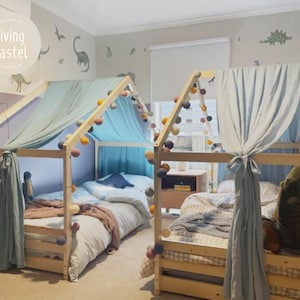 May include: Two wooden house-shaped beds with light blue draped fabric and colorful pom-pom garlands. The beds are made up with bedding and pillows. The room has dinosaur wall decals and the text "Living Pastel".