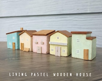 Puede incluir: Un conjunto de cinco casas de madera en miniatura en colores pastel que incluyen azul claro, crema, rosa, amarillo pálido y verde menta. Cada casa tiene un techo marrón, pequeñas ventanas cuadradas y una puerta de madera. El texto LIVING PASTEL WOODEN HOUSE es visible.