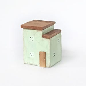 May include: A small, two-tiered house-shaped object with a pale mint green exterior and brown wooden roofs. The structure features a rectangular brown door and several small, square windows, each containing four tiny circular cutouts.