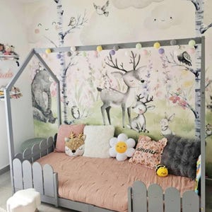May include: A gray wooden bed frame with a picket fence design, a pink comforter, and decorative pillows. The pillows include a deer-shaped pillow, a white fluffy pillow, and a flower-shaped pillow. The wall features a mural with deer and forest elements.